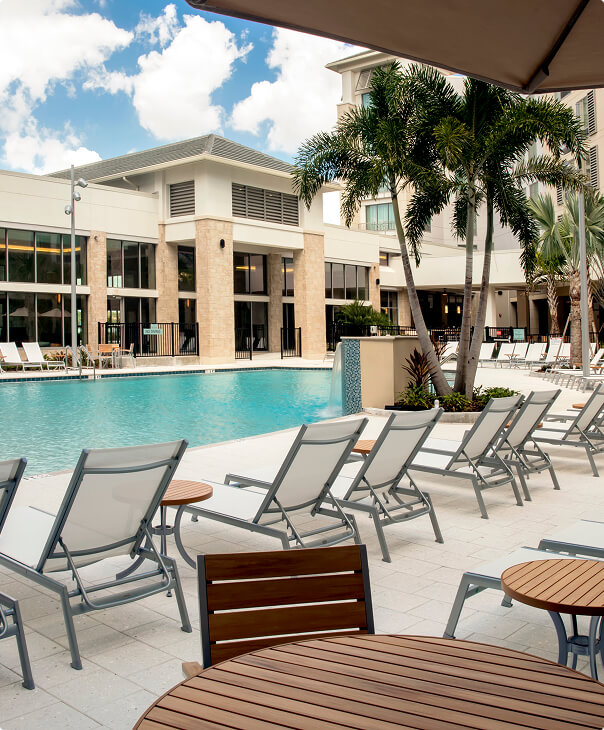 Aerial view of a poolside area with white lounge chairs and round wooden side tables on light tile flooring and a small landscaped palm tree island near the swimming pool.