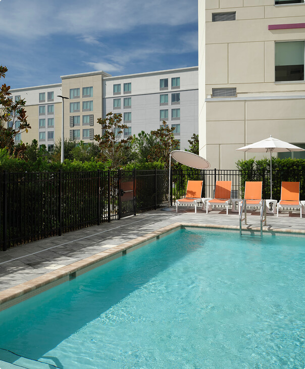 Aerial view of a poolside area with white lounge chairs and round wooden side tables on light tile flooring and a small landscaped palm tree island near the swimming pool.
