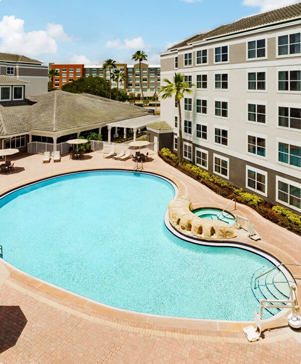 Aerial view of a poolside area with white lounge chairs and round wooden side tables on light tile flooring and a small landscaped palm tree island near the swimming pool.