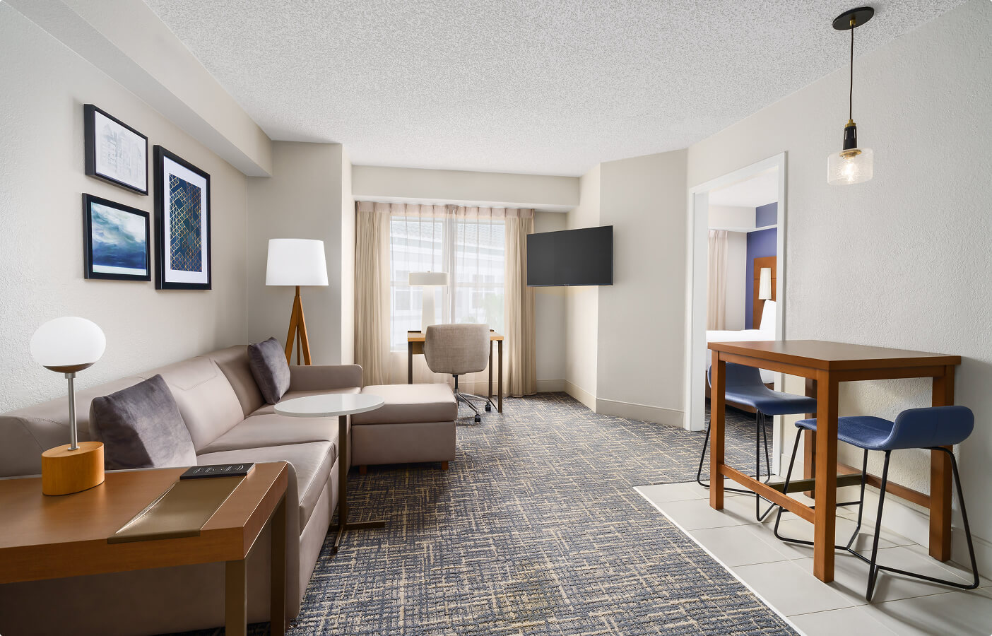 Modern hotel suite with sofa, coffee table, desk, wall-mounted TV, dining table, and two blue chairs.