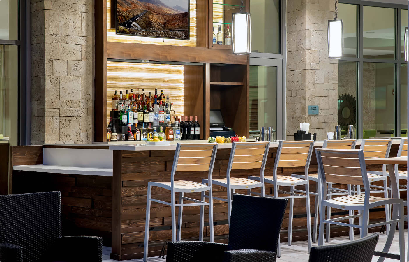 Modern indoor bar with wooden counter, high chairs, bottles, and illuminated stone wall panels.