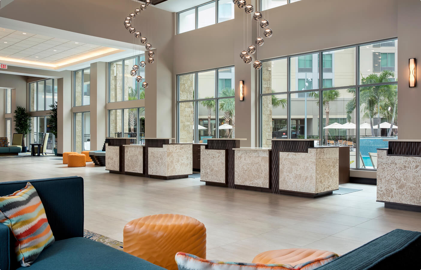 Modern hotel lobby with reception desks, large windows, hanging lights, seating, and pool view outside.