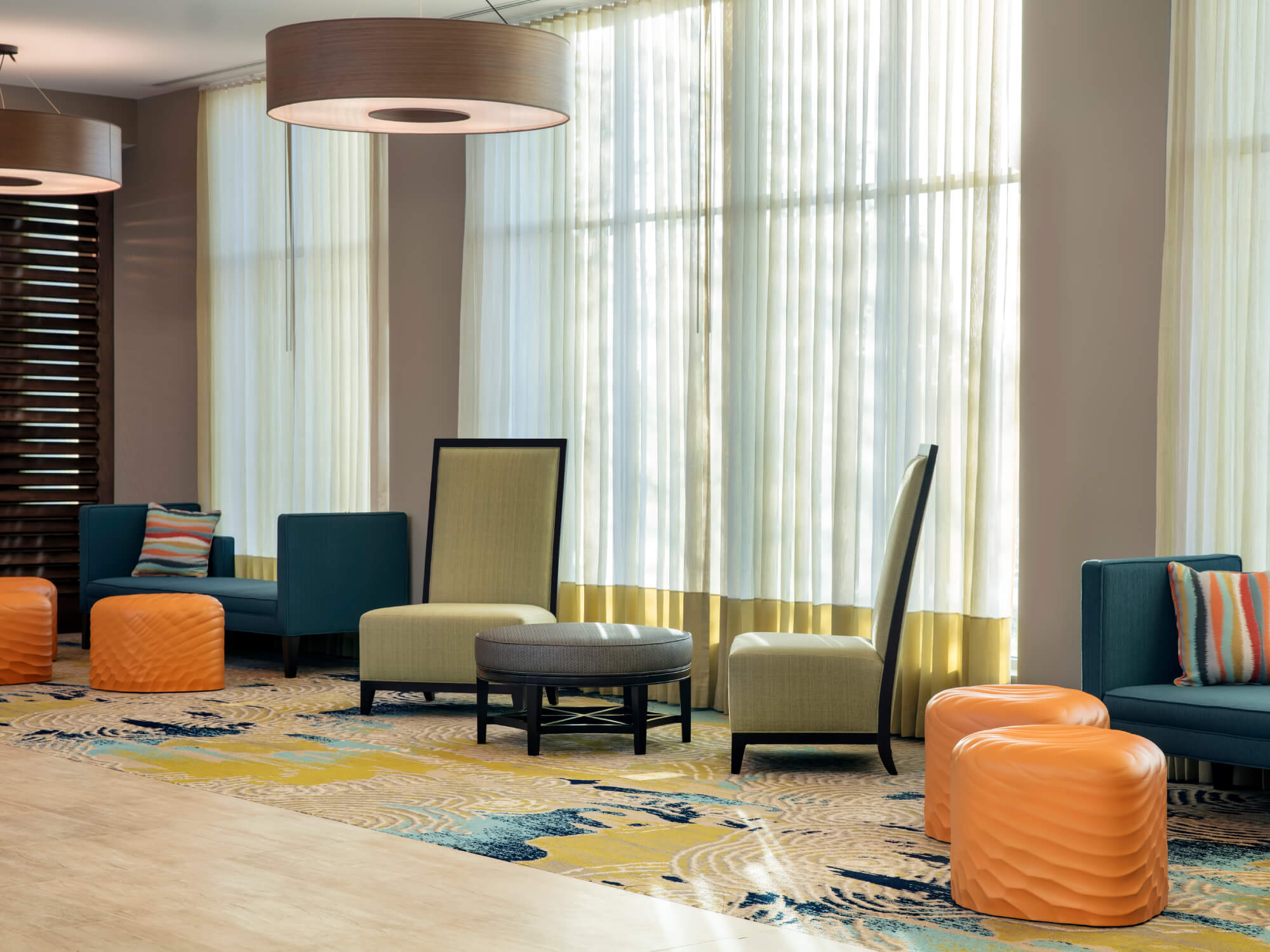 Hotel lobby with beige chairs, blue couches, orange poufs, and large windows with sheer curtains.