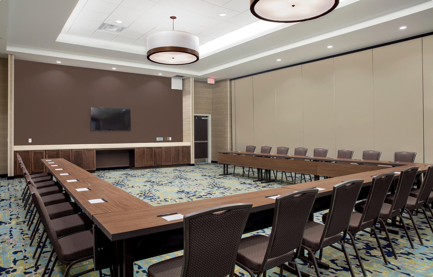 Conference room with U-shaped wooden tables, chairs, carpeted floor, and wall-mounted TV.
