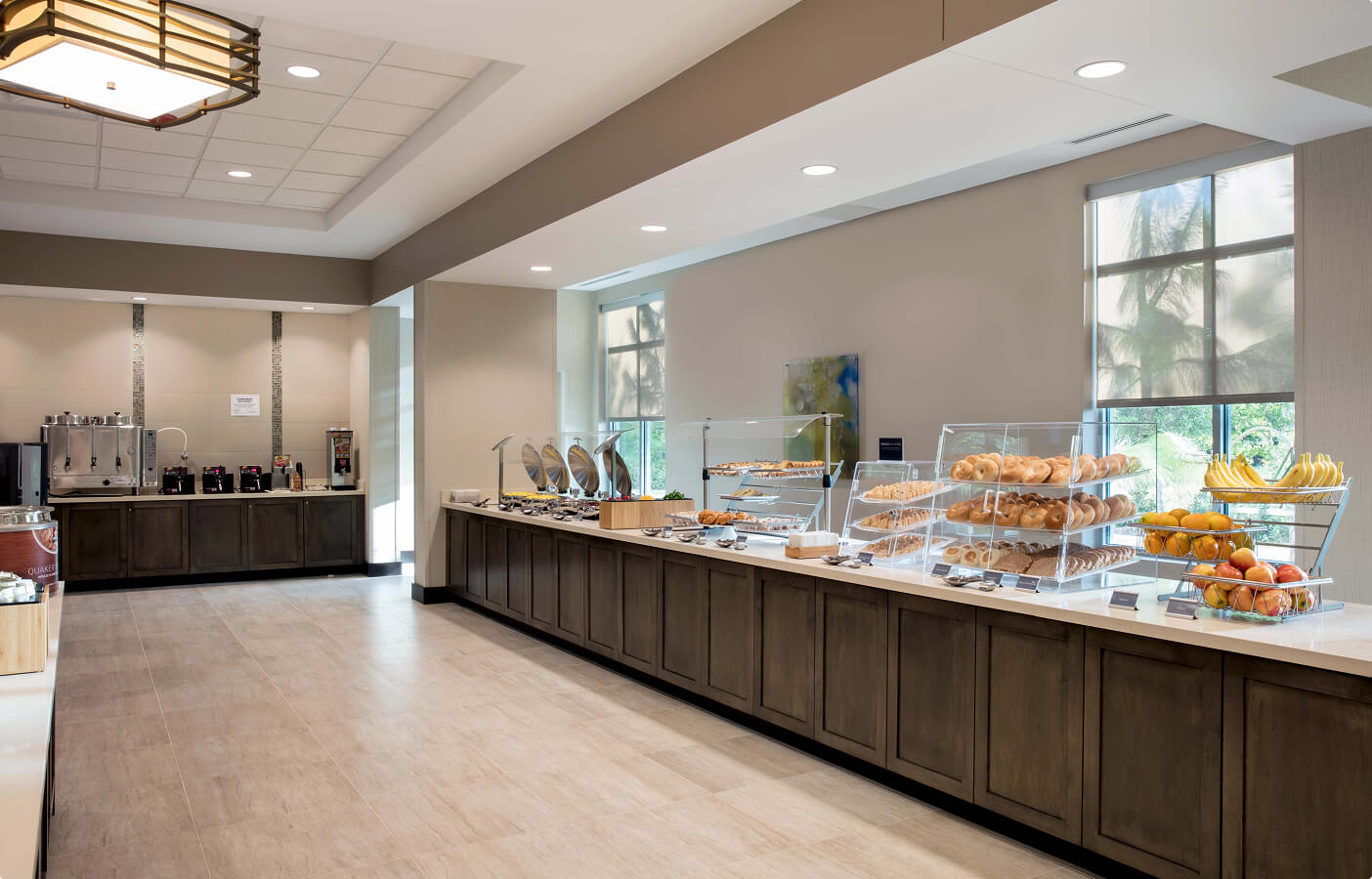 Hotel breakfast buffet with pastries, bagels, fruit, hot food trays, and beverage dispensers in a bright room.