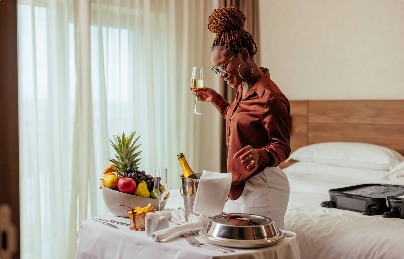 Woman with braided hair and glasses holding a glass of champagne near a hotel room service table.