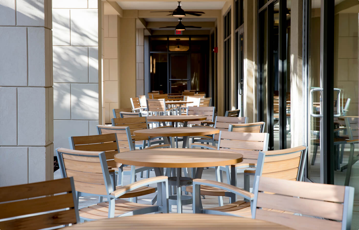 Empty outdoor seating area with wooden tables and chairs along a building corridor.