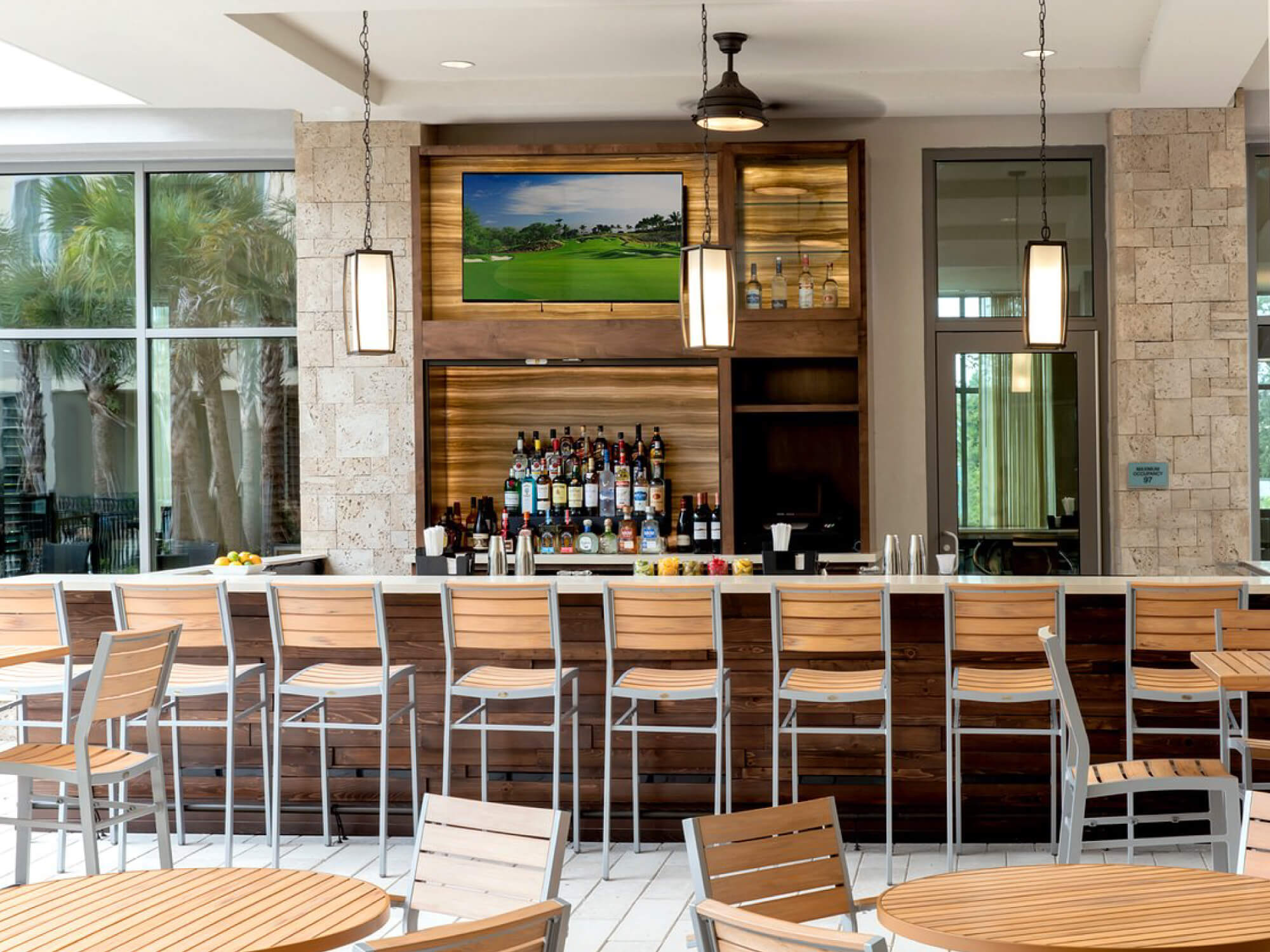 Modern bar area with wooden stools, pendant lights, bottles on shelves, and a golf course TV screen.