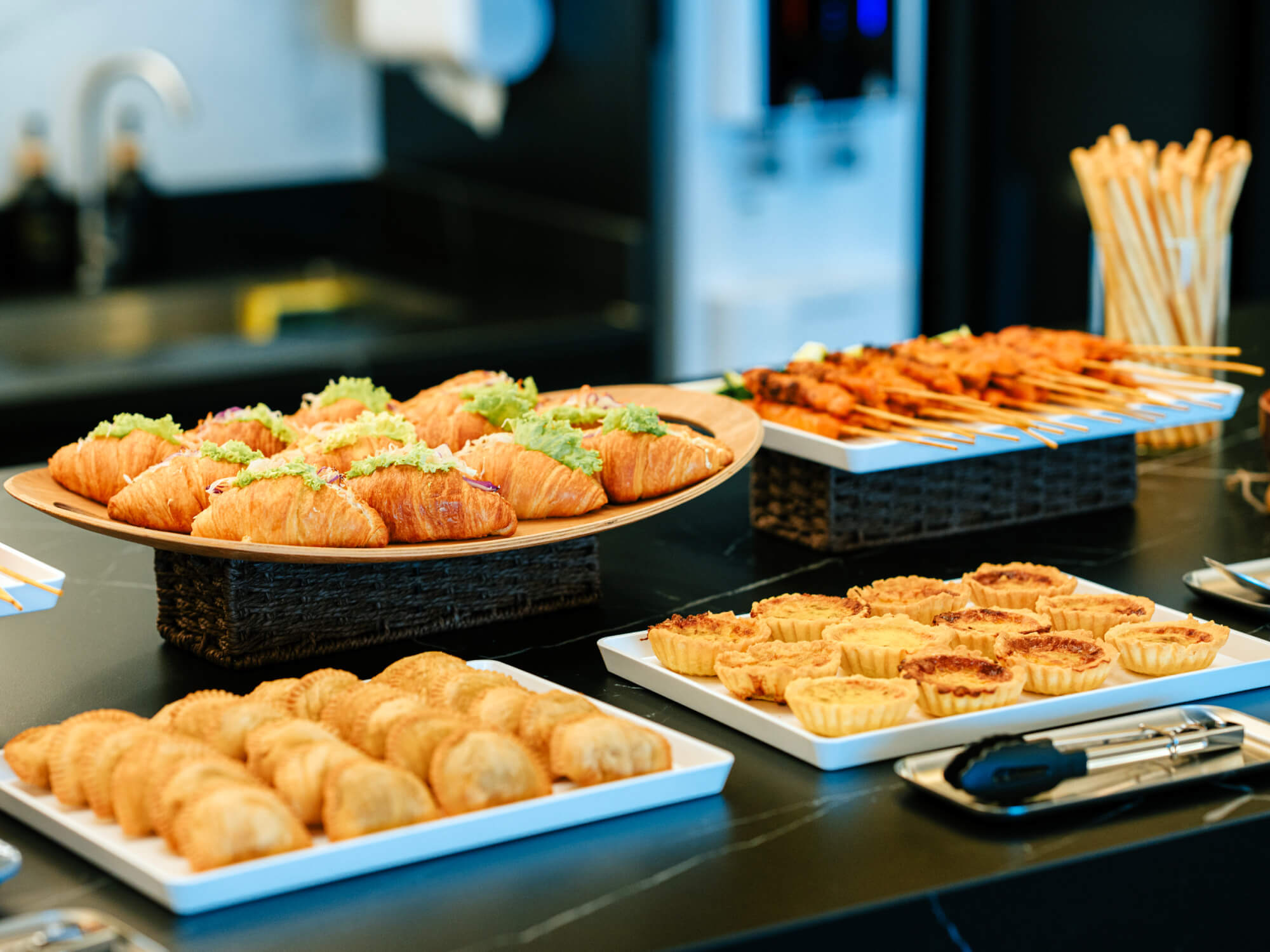 Buffet table with croissant sandwiches, skewered meat, mini tarts, and pastries on white trays.