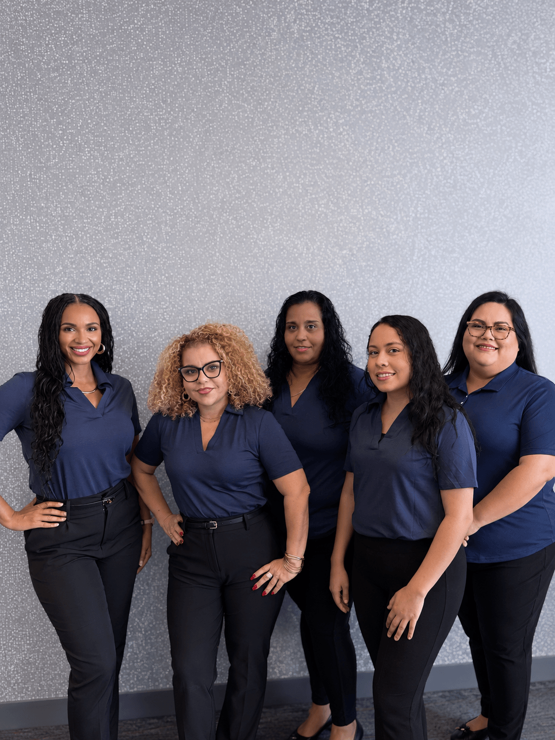 Five diverse women wearing navy shirts and black pants standing in front of a gray patterned wall.