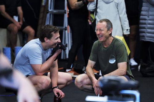 A fit individual performs a jump over dumbbells in a gym filled with spectators, showcasing athleticism and energy during a workout session.