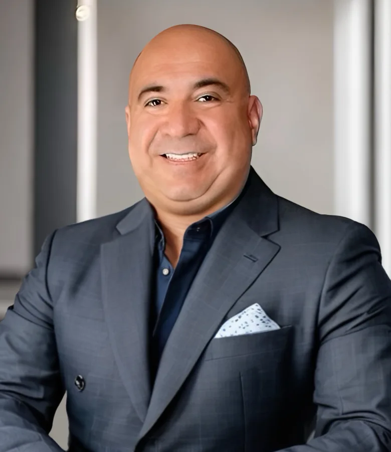 Smiling bald man in a dark blue suit jacket with a light blue patterned pocket square against a blurred background.