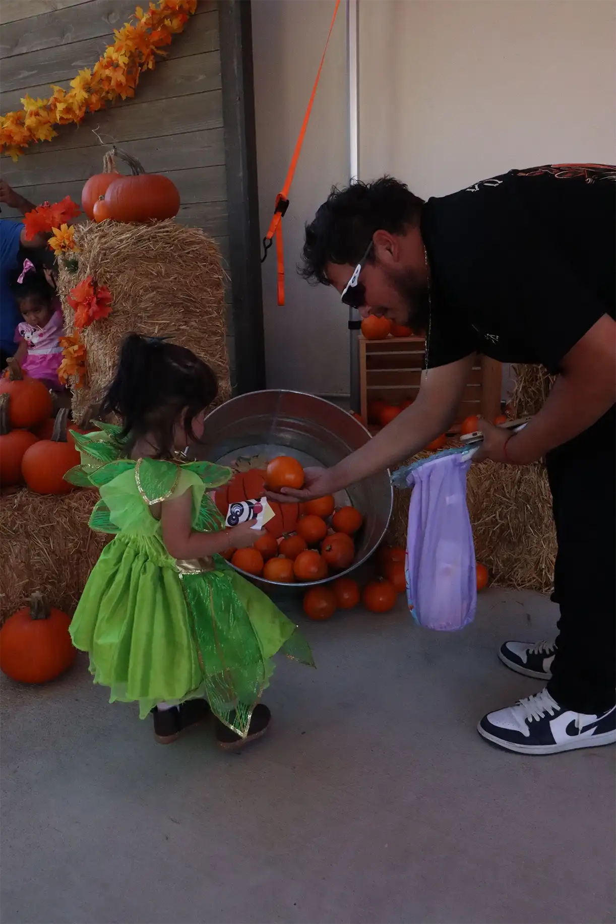 Child in green costume picking pumpkins from metal tub during Halloween celebration