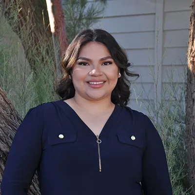 Smiling woman in navy top standing outdoors near trees and white wall