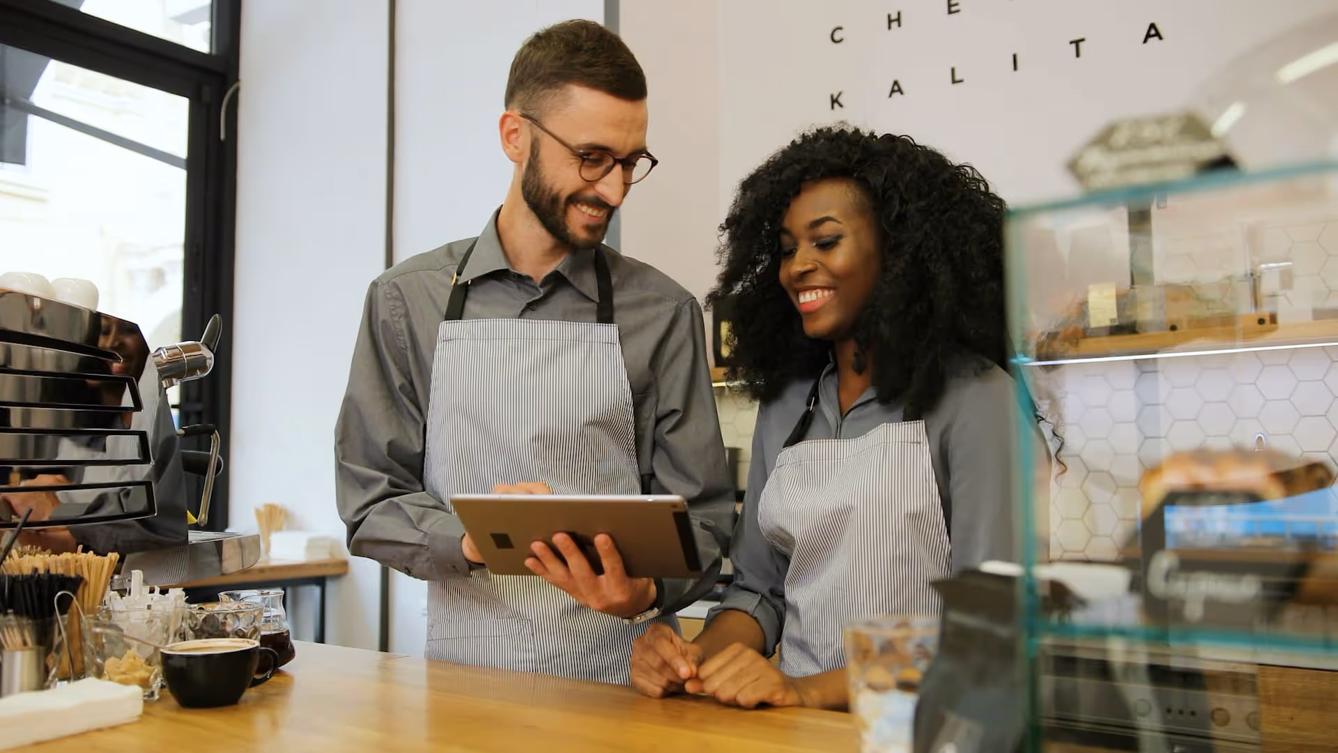 Two smiling baristas wearing aprons discuss orders while looking at a digital tablet behind a coffee counter.