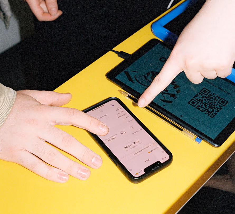 Two people at a yellow table interacting with a smartphone showing a schedule app and a tablet displaying a QR code, one hand pointing at the tablet screen.