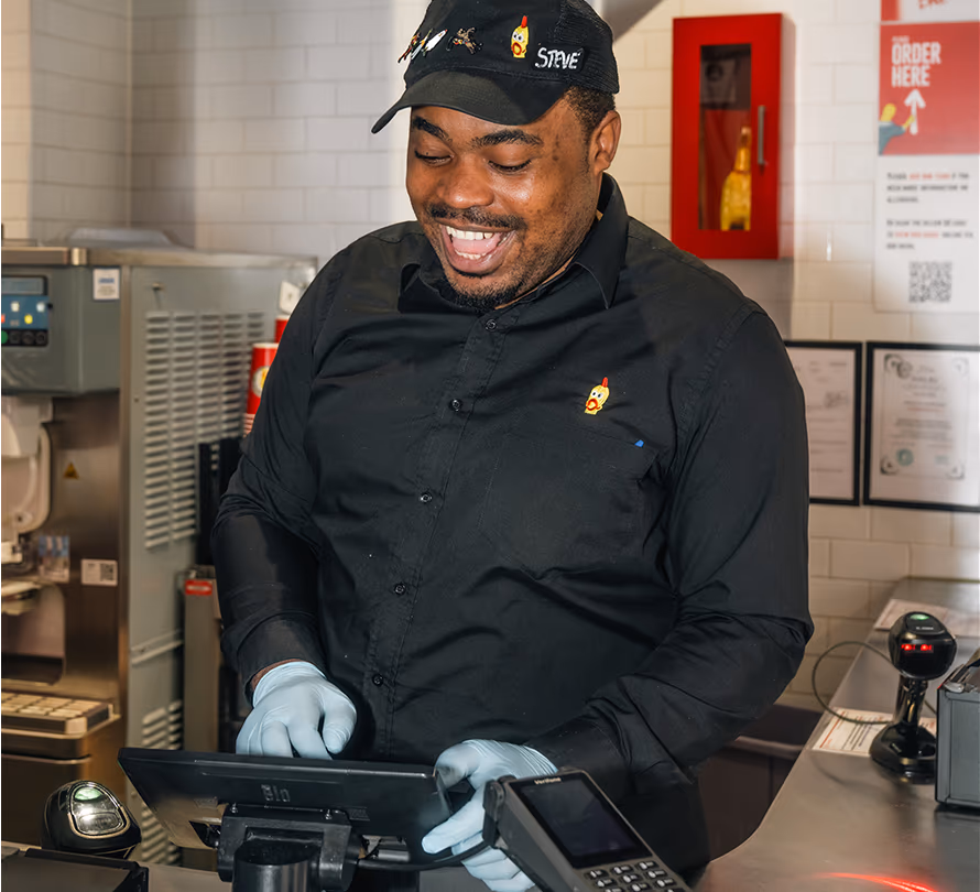 A smiling employee wearing a black cap and shirt uses a point-of-sale touchscreen at a restaurant counter.