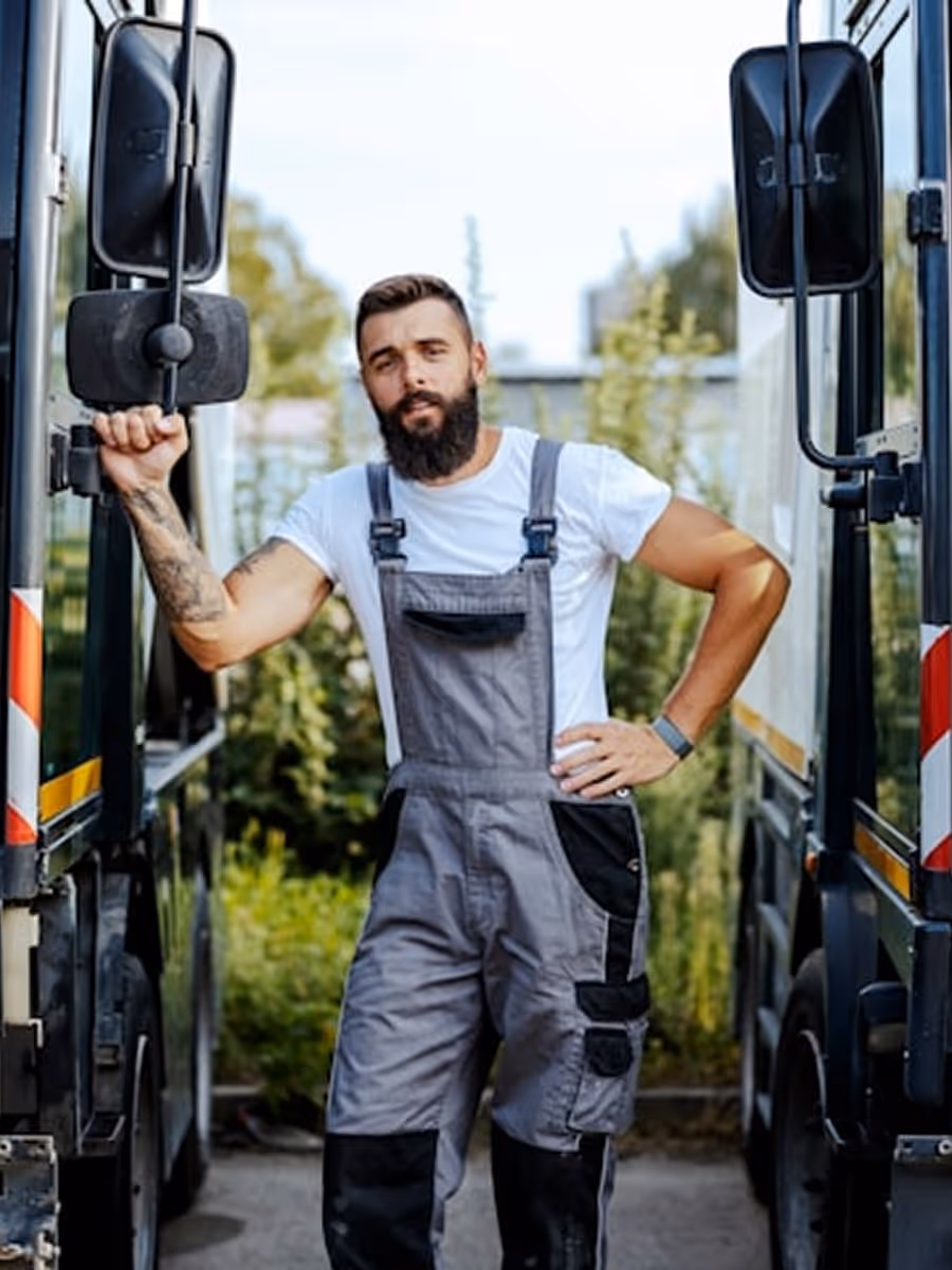 A man with a beard and overalls standing in front of a bus.