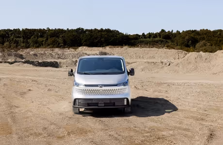 Silberner Maxus eDeliver 7 mit Allradantrieb von vorne, geparkt in sandiger Landschaft vor Baumkulisse – moderner Elektro-Transporter für anspruchsvolle Einsätze.