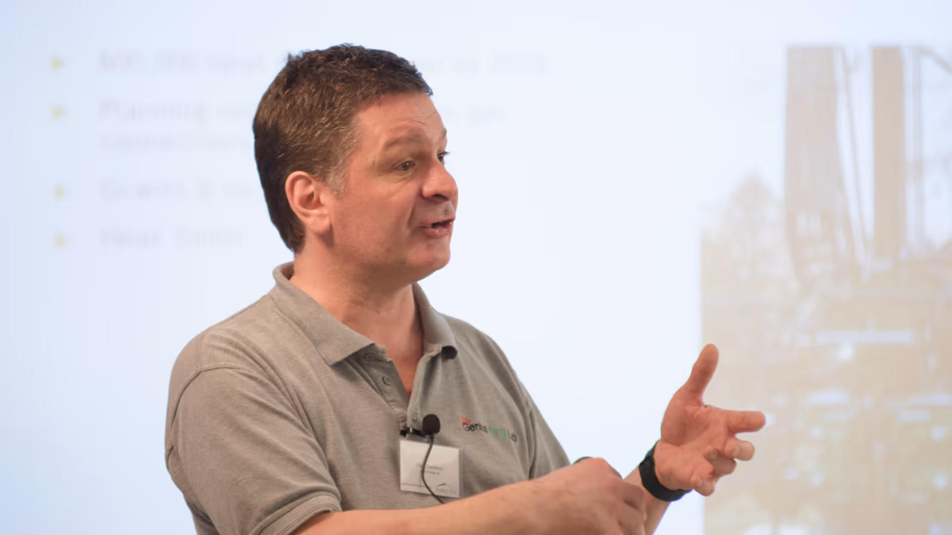Man with short dark hair wearing a gray polo shirt and speaking with hand gestures in front of a blurred presentation screen.
