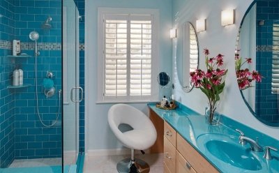 Remodeled bathroom with blue-tiled walk-in shower, blue counter tops over light-brown cabinets, and two round mirrors 