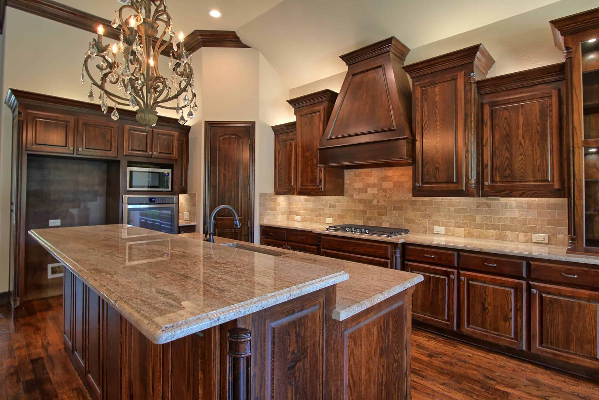 Elegant Oakmark Signature Homes kitchen with rich wood cabinetry, ornate chandelier, stone backsplash, and large granite island centerpiece.