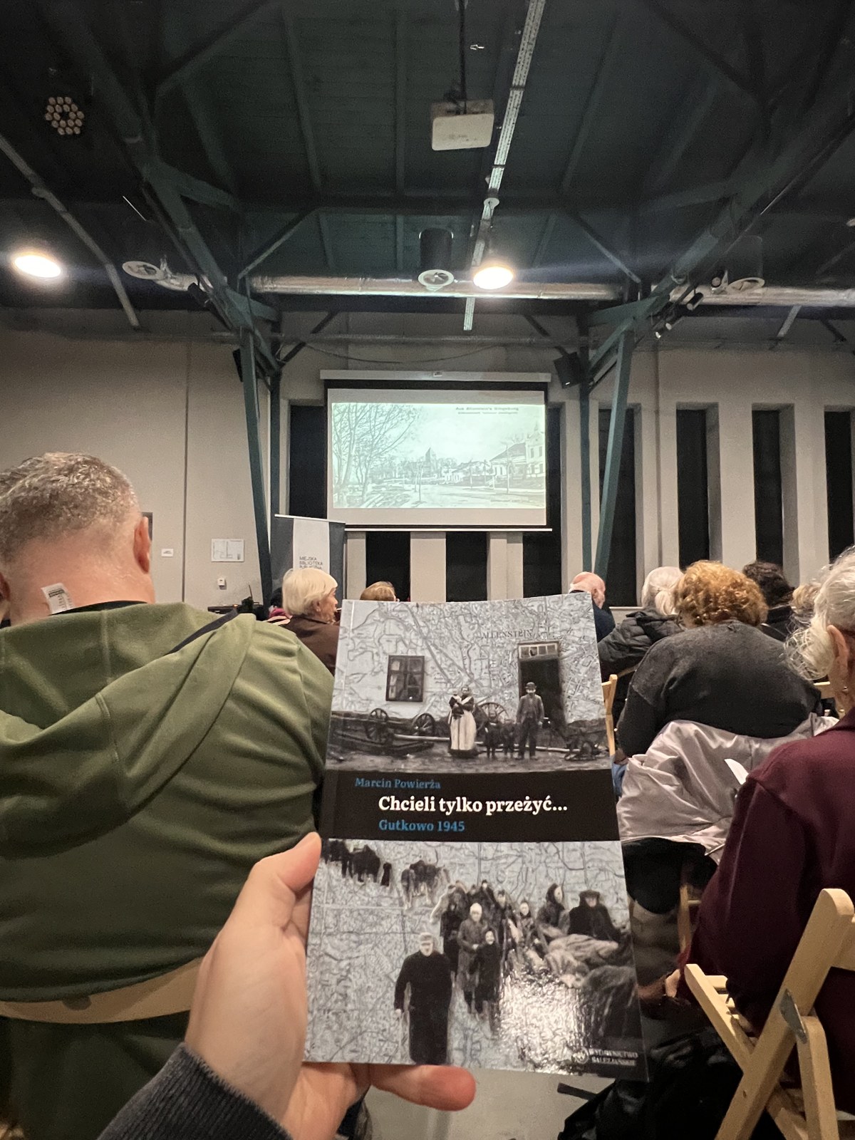 A reader holds up a copy of Chcieli tylko przezyc - Gutkowo 1945 during the book talk, an archival photograph on the screen behind
