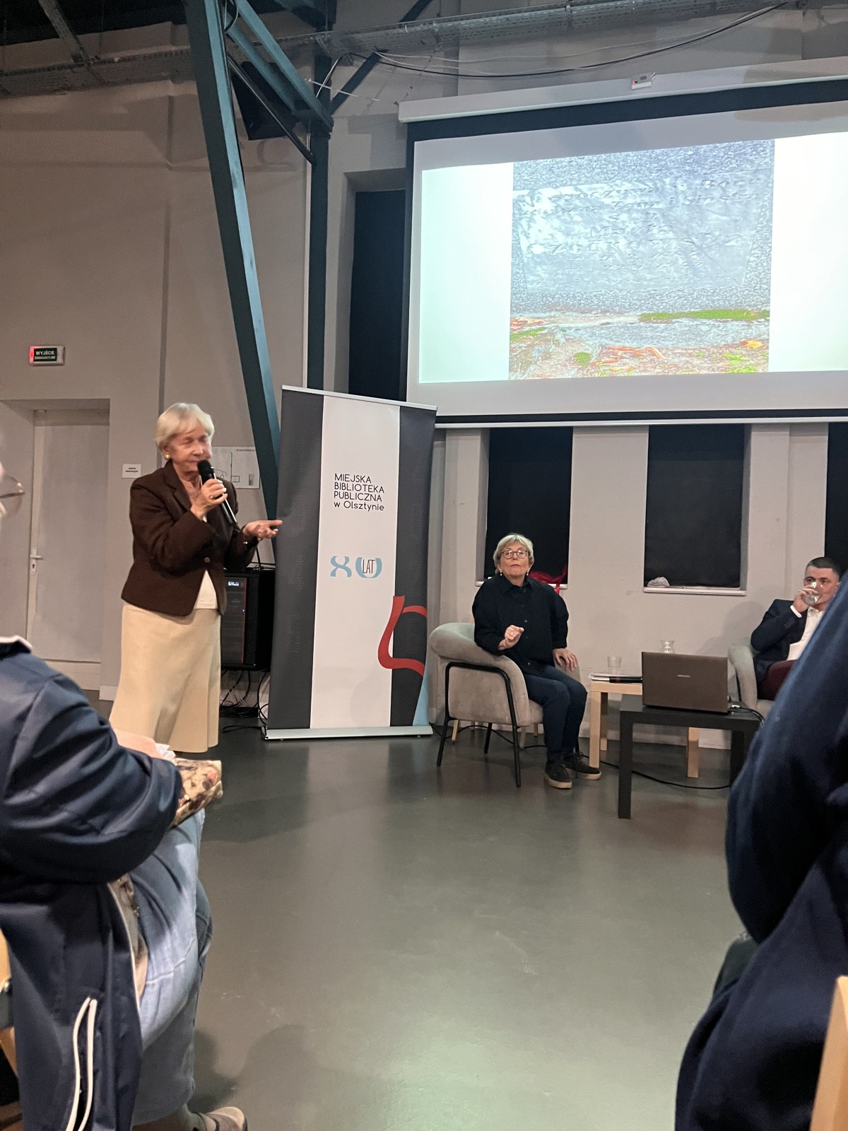 An older woman stands with a microphone to share her own memory at the end of the book talk in the Olsztyn public library