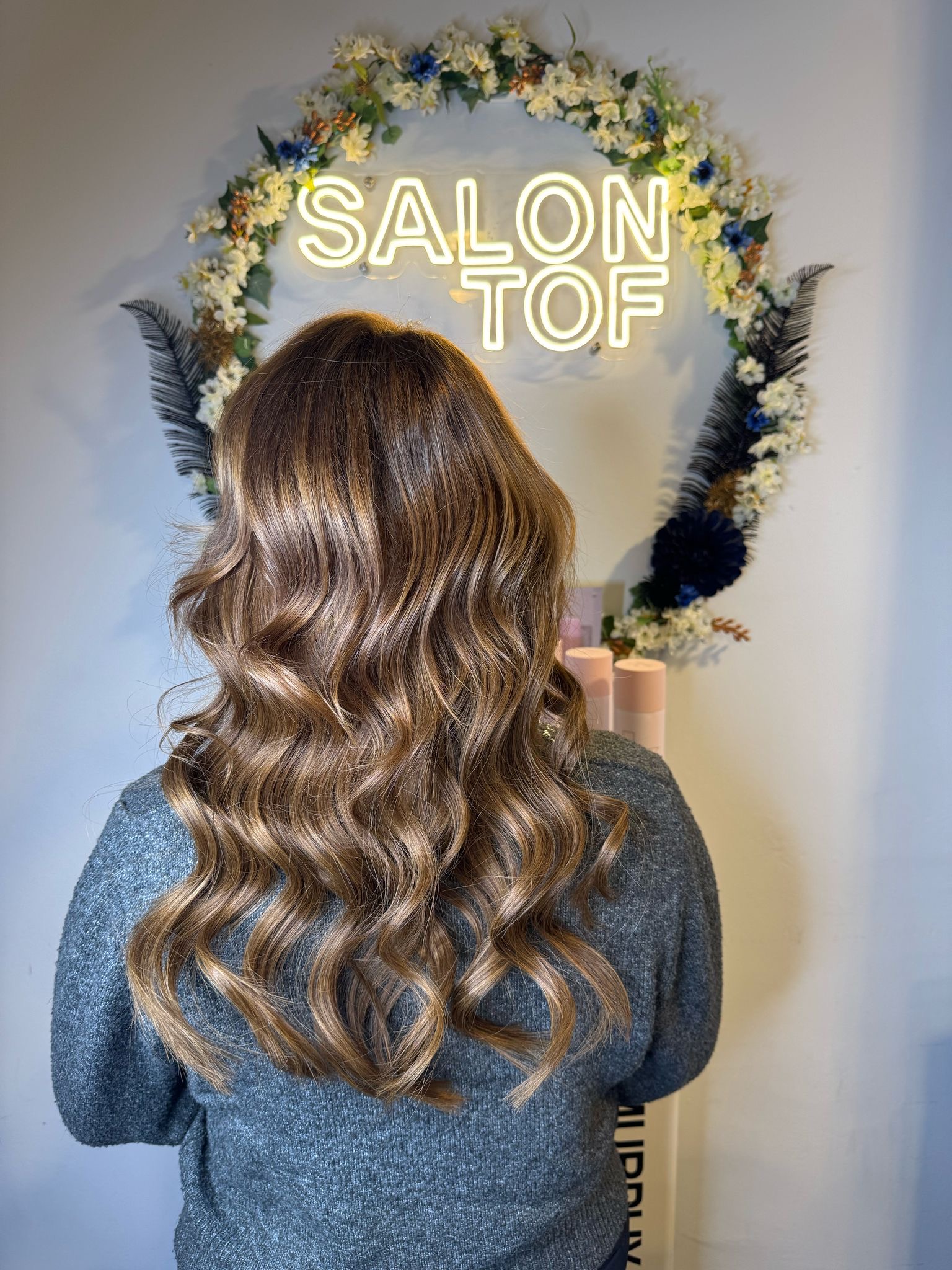 Een vrouw staat met haar rug naar de camera in een kapsalon in 's-Hertogenbosch, met mooi golvend bruin haar, voor een verlicht naambord met de tekst "SALON TOF" omgeven door een bloemenkrans.