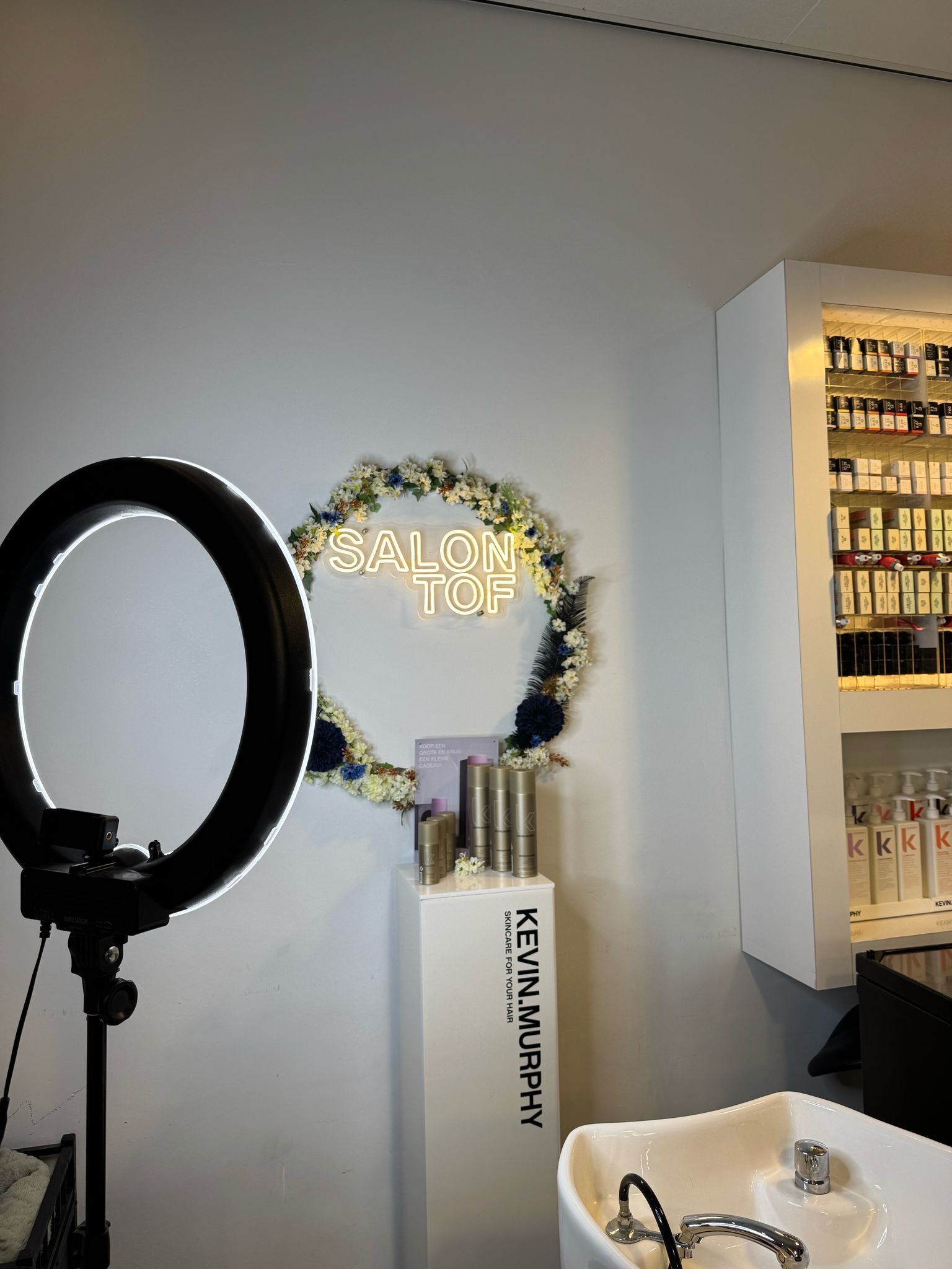 Interieur van een kapperszaak in 's-Hertogenbosch met een verlichte ringlamp, een wasbak, en een muurdecoratie dat "SALON TOF" omringd door bloemen leest.