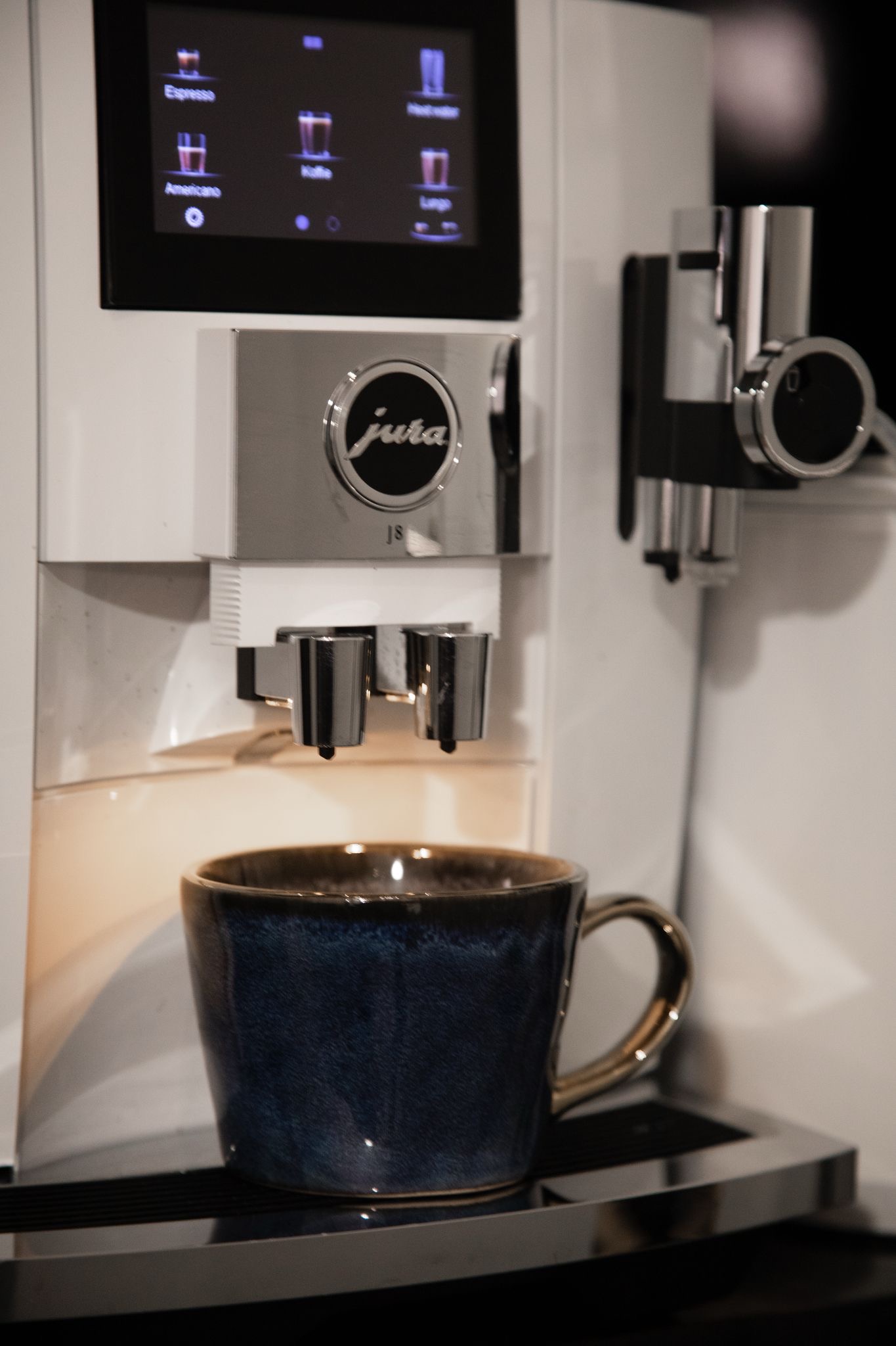 Koffiezetapparaat van het merk Jura dat verschillende koffie-opties toont op een digitaal display, met een blauwe mok eronder waar koffie in geschonken wordt.