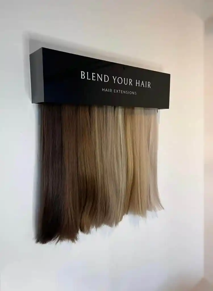 Een muurdisplay in een kapsalon toont een scala aan haarextensies in diverse blonde en bruine tinten met daarboven een zwart bord met de tekst 'BLEND YOUR HAIR HAIR EXTENSIONS'.