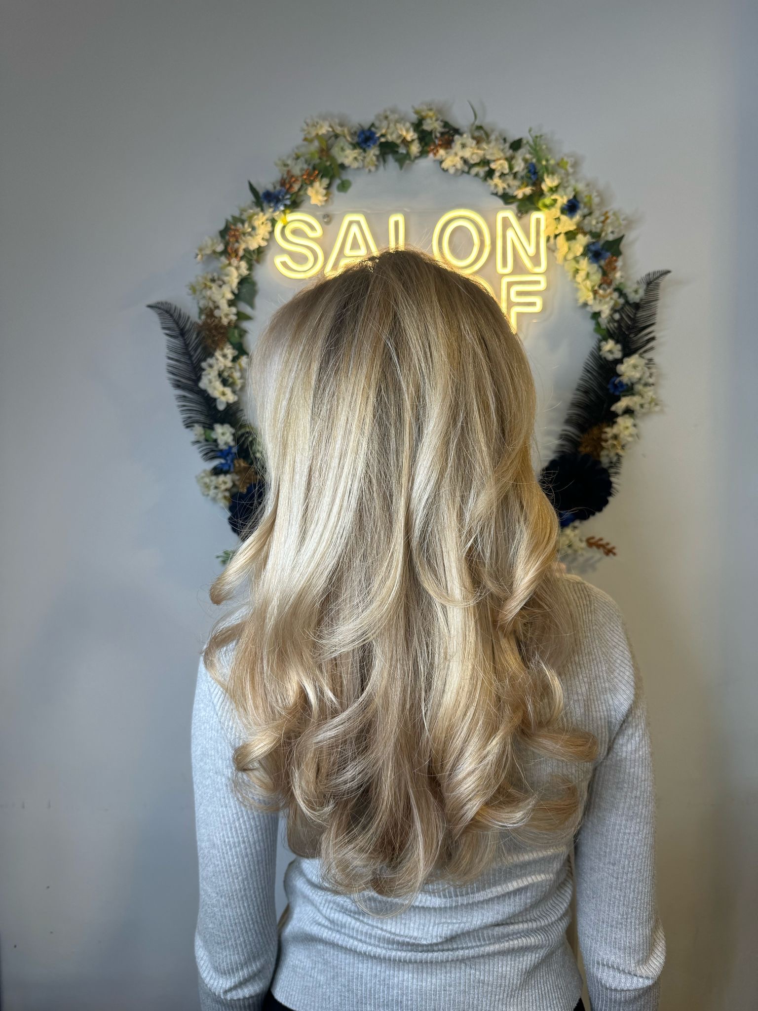 Een vrouw toont haar pas gekapte blonde haar in zachte golven, met haar rug naar de camera, tegen de achtergrond van een verlicht 'SALON' bord omringd door een bloemenkrans in een kapsalon in 's-Hertogenbosch.