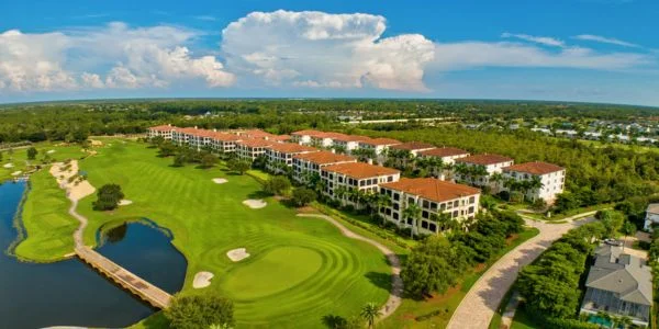 Aerial view of a golf course with a pond, sand traps, and a row of red-roofed residential buildings surrounded by greenery under a blue sky with clouds.