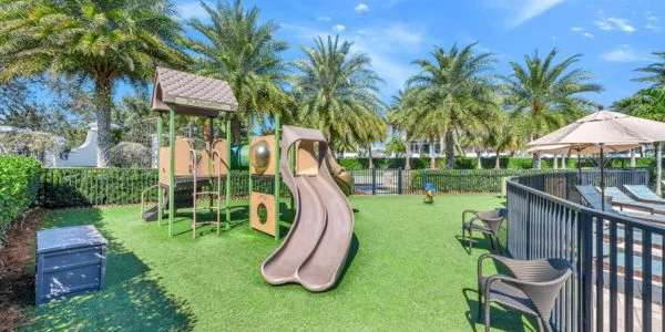 Children's playground with slides and climbing structures on artificial grass surrounded by palm trees and a fenced pool area with lounge chairs under umbrellas.
