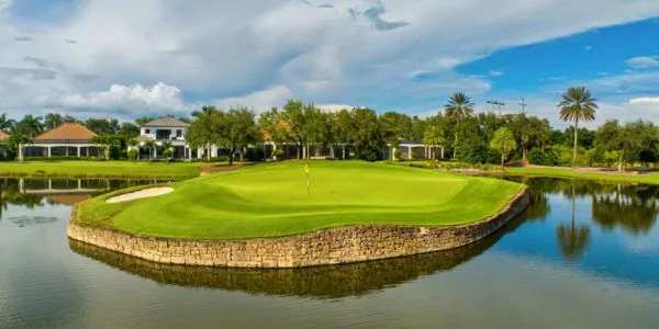 Golf green on an island surrounded by water with stone retaining walls and houses, palm trees, and a partly cloudy sky in the background.
