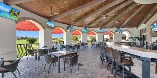 Covered outdoor seating area with tables, chairs, ceiling fans, and mounted TVs overlooking a green landscape.