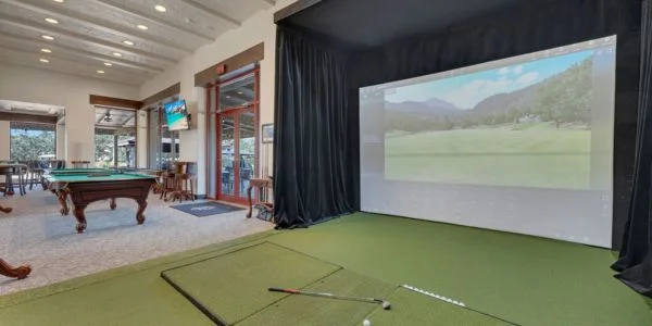 Indoor golf simulator area with a green putting mat, golf clubs, balls, and a large screen showing a golf course, adjacent to a room with a pool table and seating.