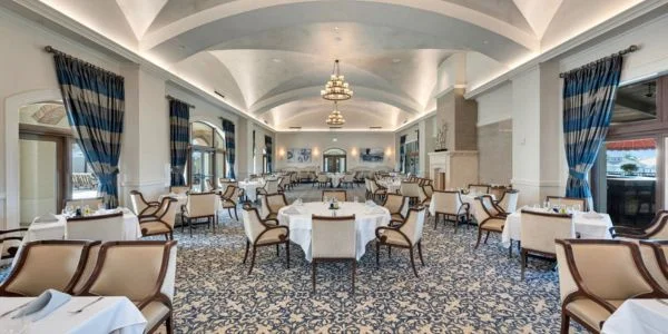 Elegant dining room with arched ceiling, chandeliers, patterned carpet, white tablecloths, and beige upholstered chairs.