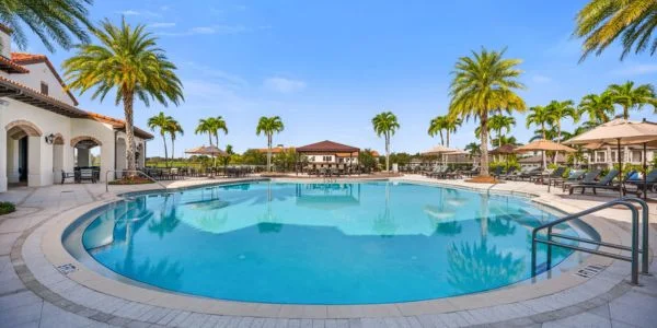 Round swimming pool surrounded by palm trees, lounge chairs, and a Mediterranean-style building under a clear blue sky.