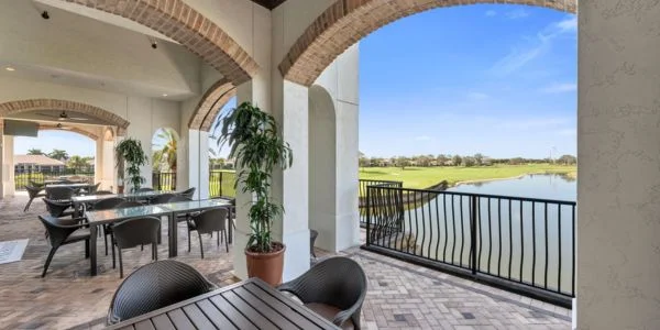 Outdoor covered patio with dining tables and chairs overlooking a lake and green landscape under a clear blue sky.