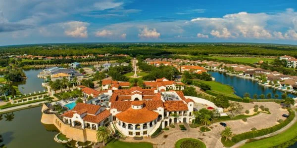 Aerial view of a lakeside residential community with Mediterranean-style buildings featuring red-tiled roofs surrounded by water and greenery under a partly cloudy blue sky.