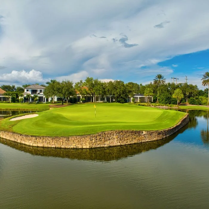 Golf green surrounded by a stone retaining wall and water with houses and trees in the background under a partly cloudy sky.