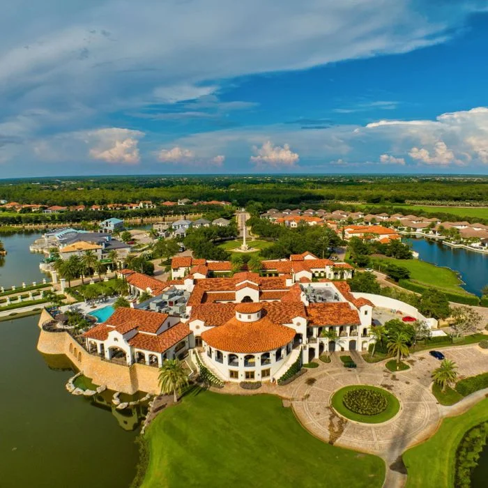 Aerial view of a large Mediterranean-style building with red-tiled roofs surrounded by water, green lawns, and residential houses under a partly cloudy blue sky.