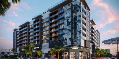 Modern multi-story apartment building with balconies and artistic mural on the corner, surrounded by palm trees under a colorful sunset sky.