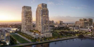 Two tall residential buildings by a waterfront with surrounding greenery and cityscape at sunset.