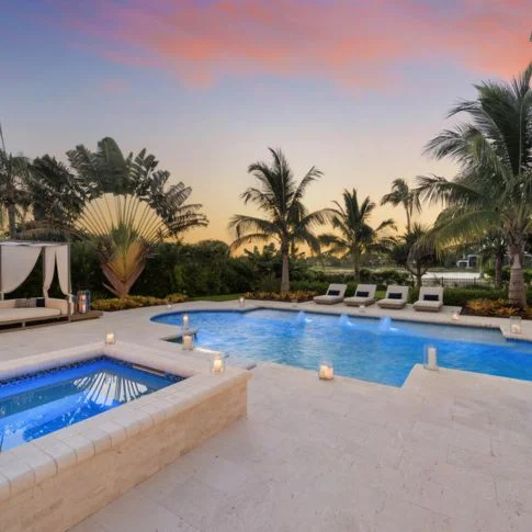 Large white luxury house with a landscaped front yard, palm trees, and a curved driveway at sunset.