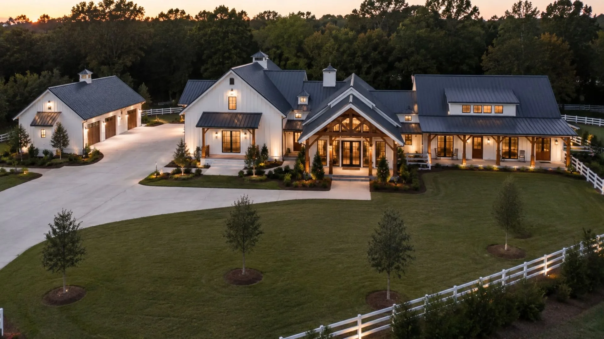 Large modern farmhouse with white siding, dark roof, lit porch, garage, and landscaped lawn at dusk.