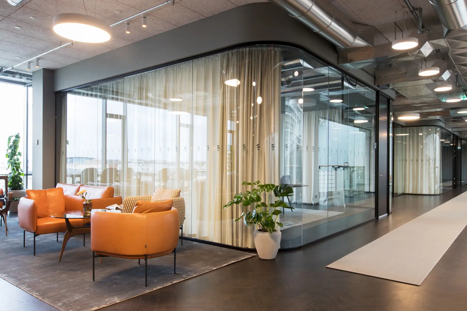Moderne kontorområder med oransje skinnstoler og mønstret lenestol rundt et lavt glassbord på et grått teppe ved et glassinnelukket møterom med beige gardiner og planter i potter.