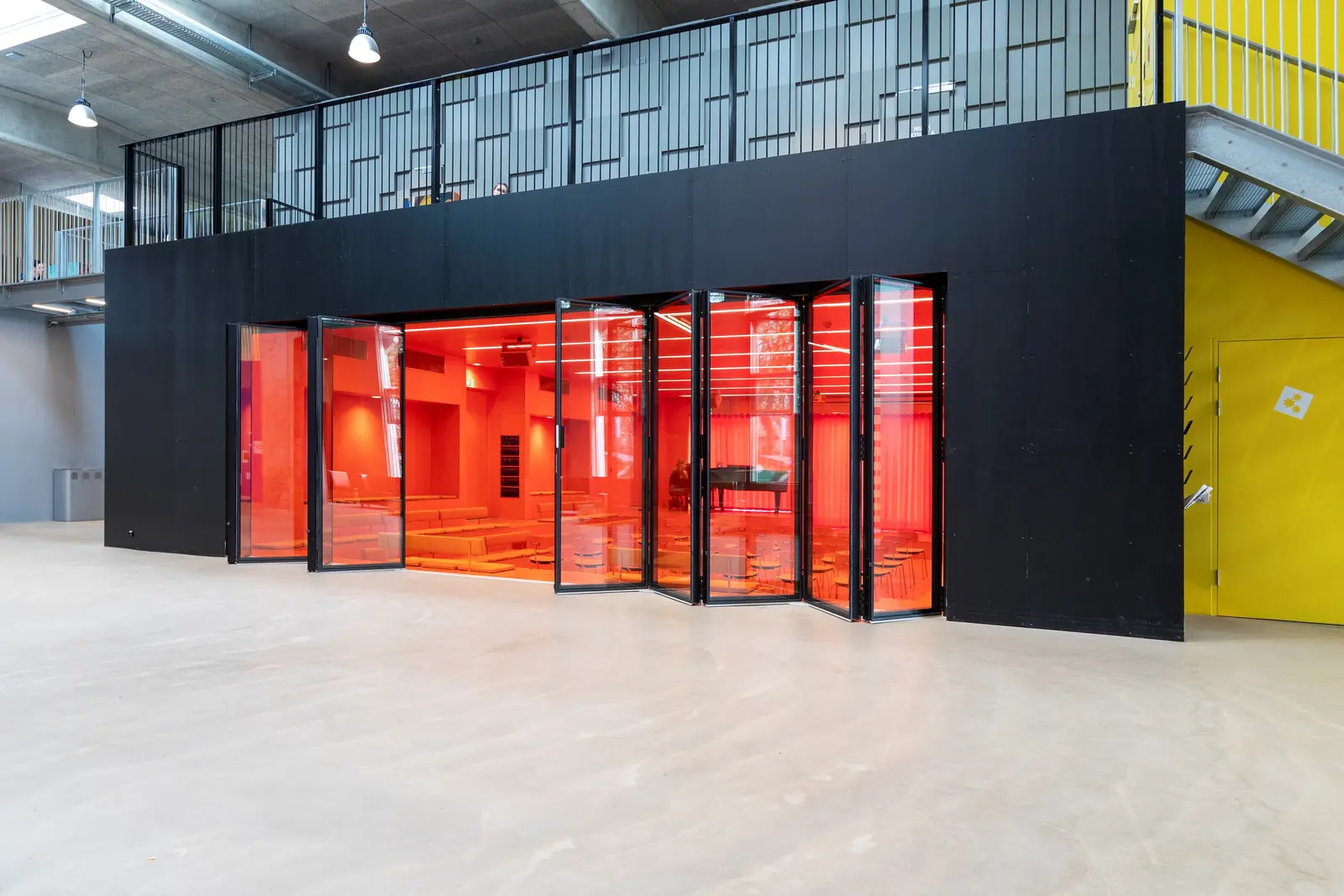Moderne rom med svarte vegger og gulnet glattt gulv, et område med store foldbare glassdører i oransjerødt glass som åpner til et interiør med sittebenker og en flygel i bakgrunnen.